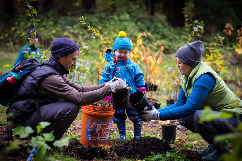 two adults plant native species while a child watches, smiling