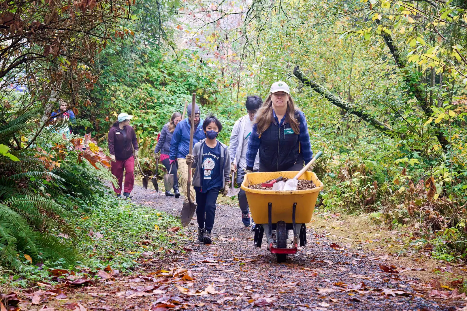 A group of people walking a trail. A woman leads the group pushing as wheelbarrow.