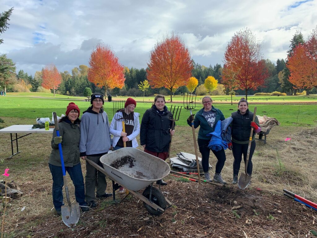 Six people posing around a wheelbarrow holding shovels and pitchforks. A field and trees are in the background.